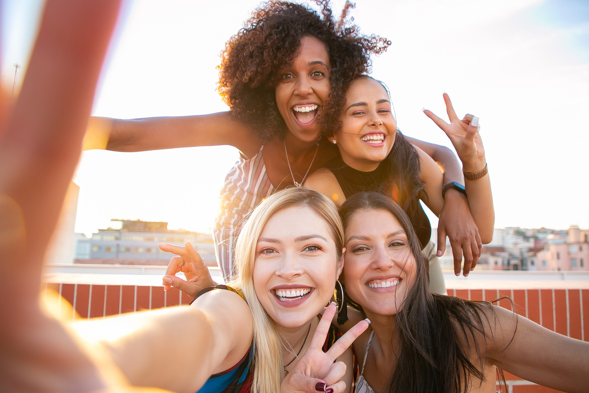 Cheerful young diverse women showing V sign while taking selfie on rooftop